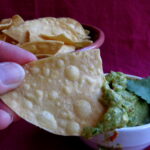 Closeup of a hand dipping a tortilla chip into Chile Avocado Sauce. Sauce is in a white bowl on top of a red cloth. There is a bowl of tortilla chips in background.