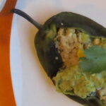 Close up of Quinoa-Stuffed Poblano Peppers with Chile Avocado Sauce spread over middle plus a sprig of cilantro. Pepper is on a white plate on top of an orange cloth.