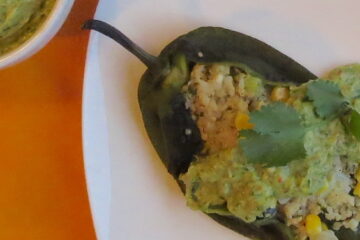 Close up of Quinoa-Stuffed Poblano Peppers with Chile Avocado Sauce spread over middle plus a sprig of cilantro. Pepper is on a white plate on top of an orange cloth.