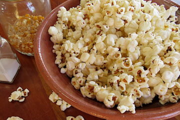 Close up of popped corn (popcorn) in a clay bowl on top of a table sprinkled with popcorn.